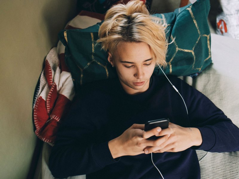 Teenager liegt auf dem Bett, das Handy in der Hand, Kopfhörer im Ohr.