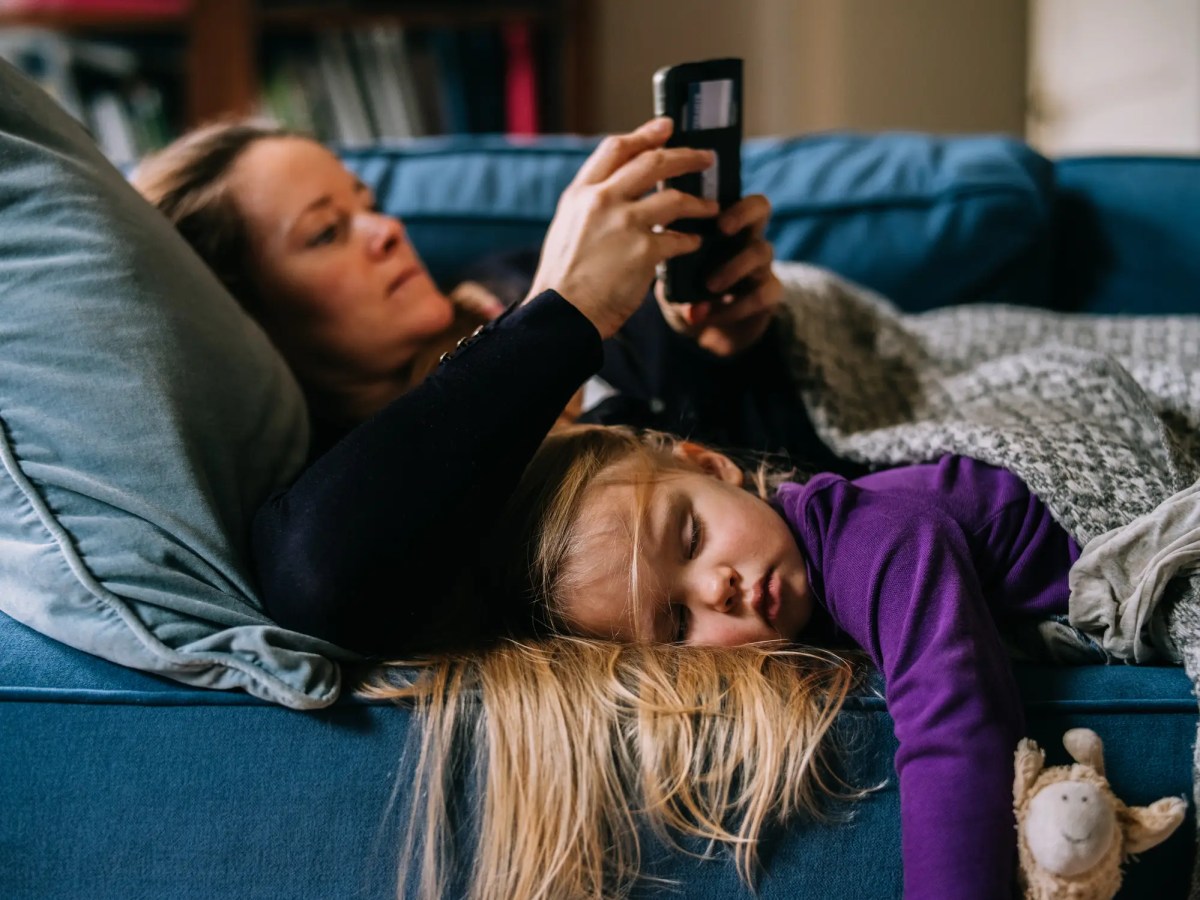Mutter liegt auf der Couch und benutzt ihr Handy. Tochter liegt daneben und schl&auml;ft