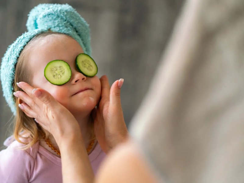 Kind mit Handtuch im Haar und Gurkenscheiben auf den Augen wird im Gesicht eingecremt.