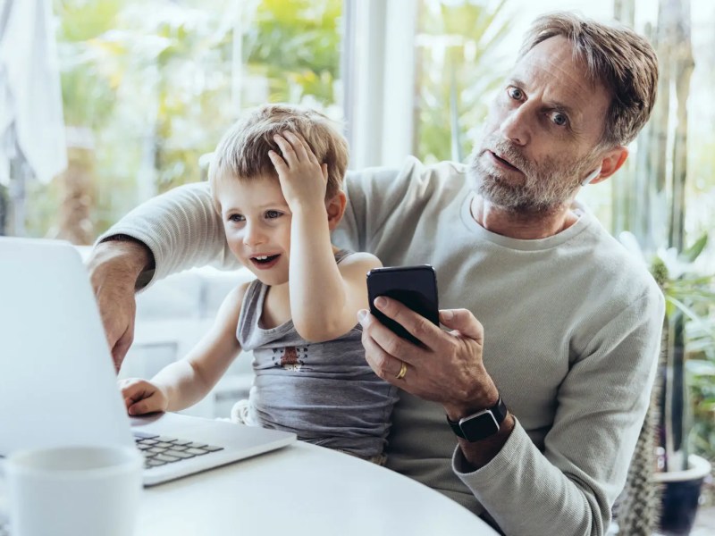 Vater mit Handy in der Hand schaut erschrocken und hat seinen Sohn auf dem Scho&szlig;, der etwas am Laptop macht.