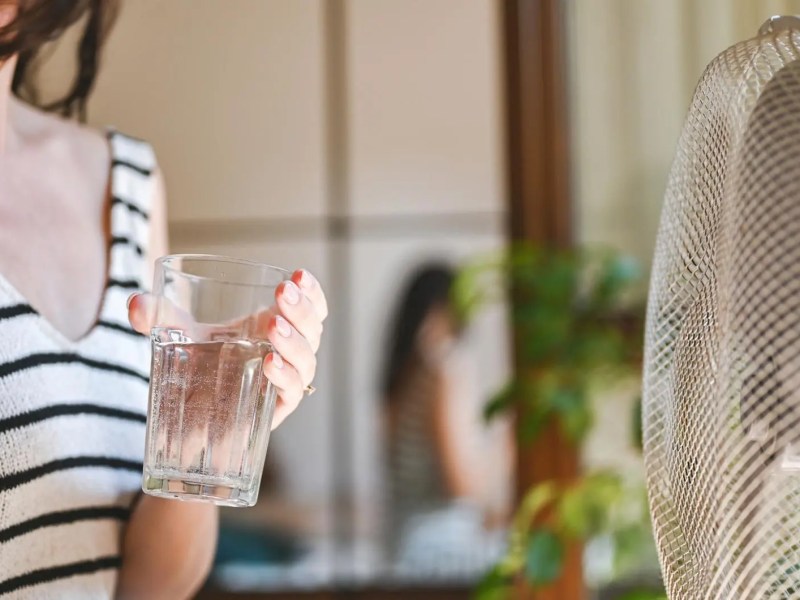 Frau mit einem Glas Wasser k&uuml;hlt sich vor dem Ventilator ab.