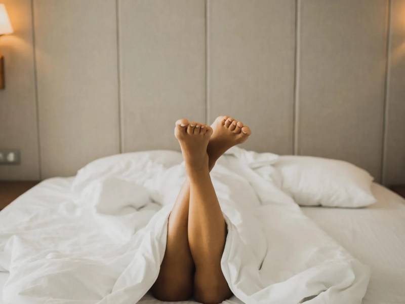 Frau liegt in einem Hotelbett, nackte Beine gucken unter der Decke hervor