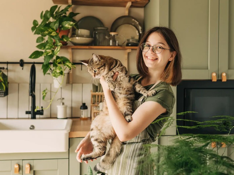 Teenagermädchen steht lachend in der Küche und hält eine Katze auf dem arm.