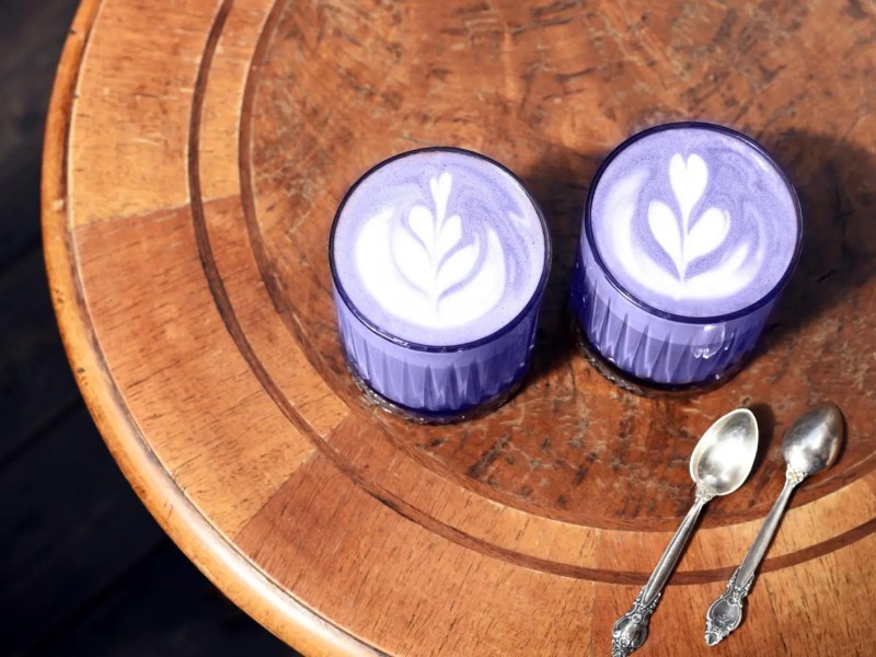 Zwei Ube Latte in violetter Farbe auf einem Holztisch mit zwei silbernen Löffeln.