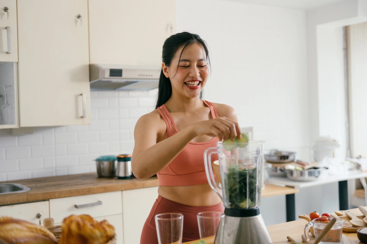 Junge Frau steht in der Küch eund pürriert sich einen gesunden, grünen Smoothie mit Gemüse.
