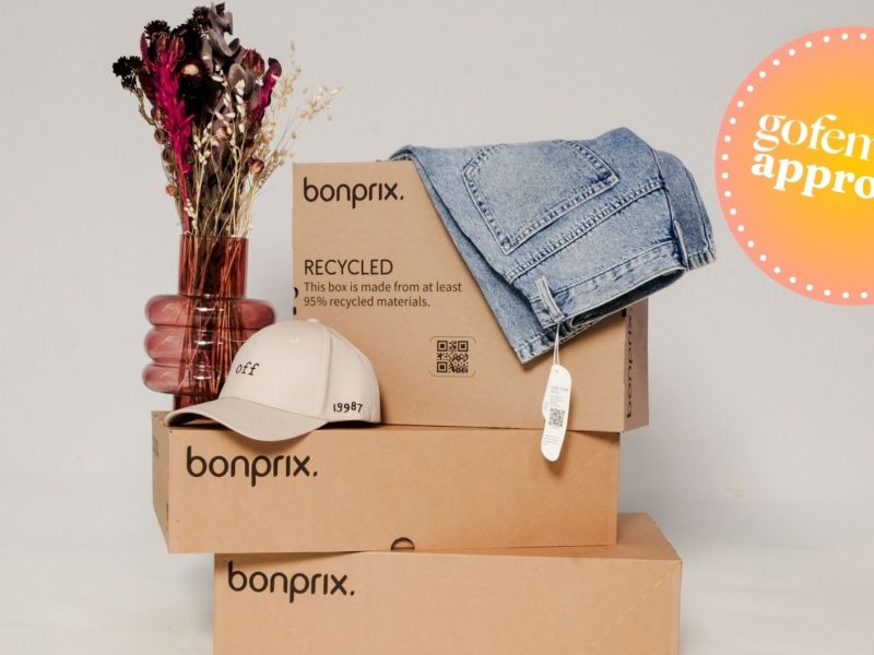 bonprix-Kisten gestapelt sowie eine Vase, eine Cap und eine Jeans.