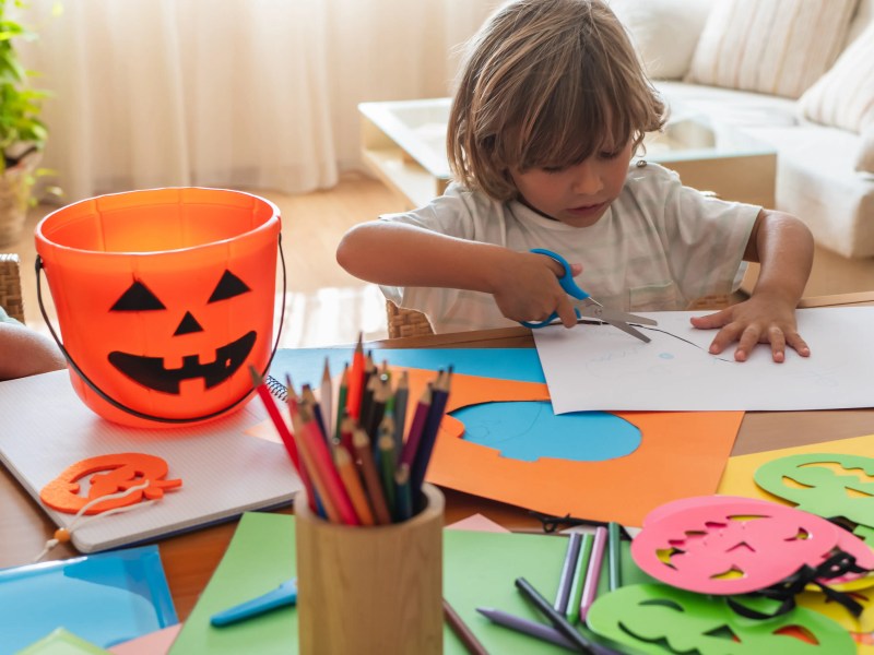 Kleinkind sitzt am Tisch und schneidet mit einer Schere Papier aus. Daneben steht ein Eimer als Halloween-K&uuml;rbis-Gesicht